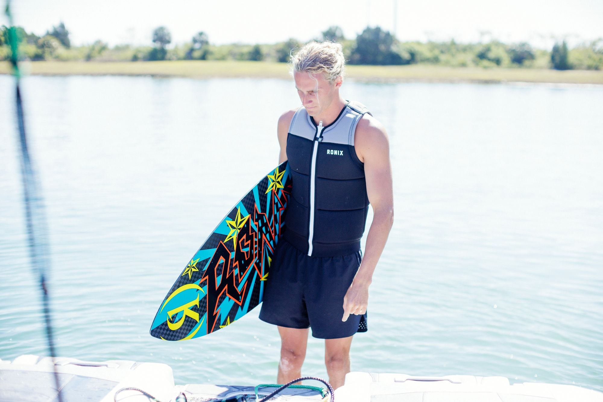 A person stands on a dock by the water, wearing the Ronix 2025 Presidente CE Approved Impact Vest and shorts, while holding a colorful wakeboard.