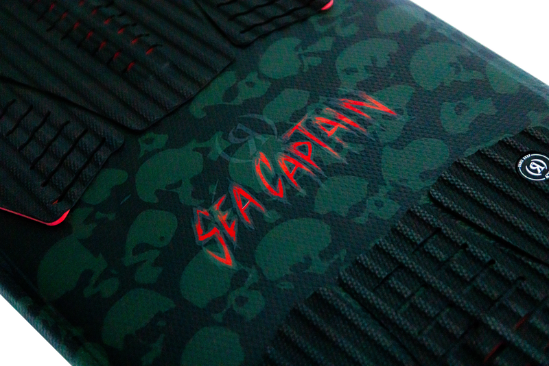 Close-up of the Ronix Sea Captain Wakesurf Board 2026 by Ronix, featuring a dark traction pad with subtle glow-in-the-dark skulls and bold red “Sea Captain” lettering.