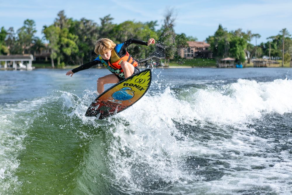 Wearing a life vest, a person rides the Ronix Sonic Fish Wakesurf Board 2026 - 3'9" across a lake, with trees and houses in the background under a clear sky.