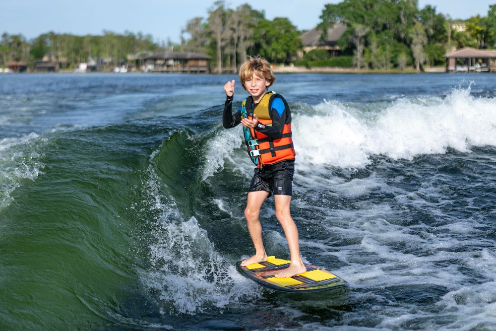 A young boy in a life jacket and board shorts rides a wave on the Ronix Sonic Fish Wakesurf Board 2026 - 3'9", smiling and giving a thumbs-up, with water splashing around and trees and houses in the background.
