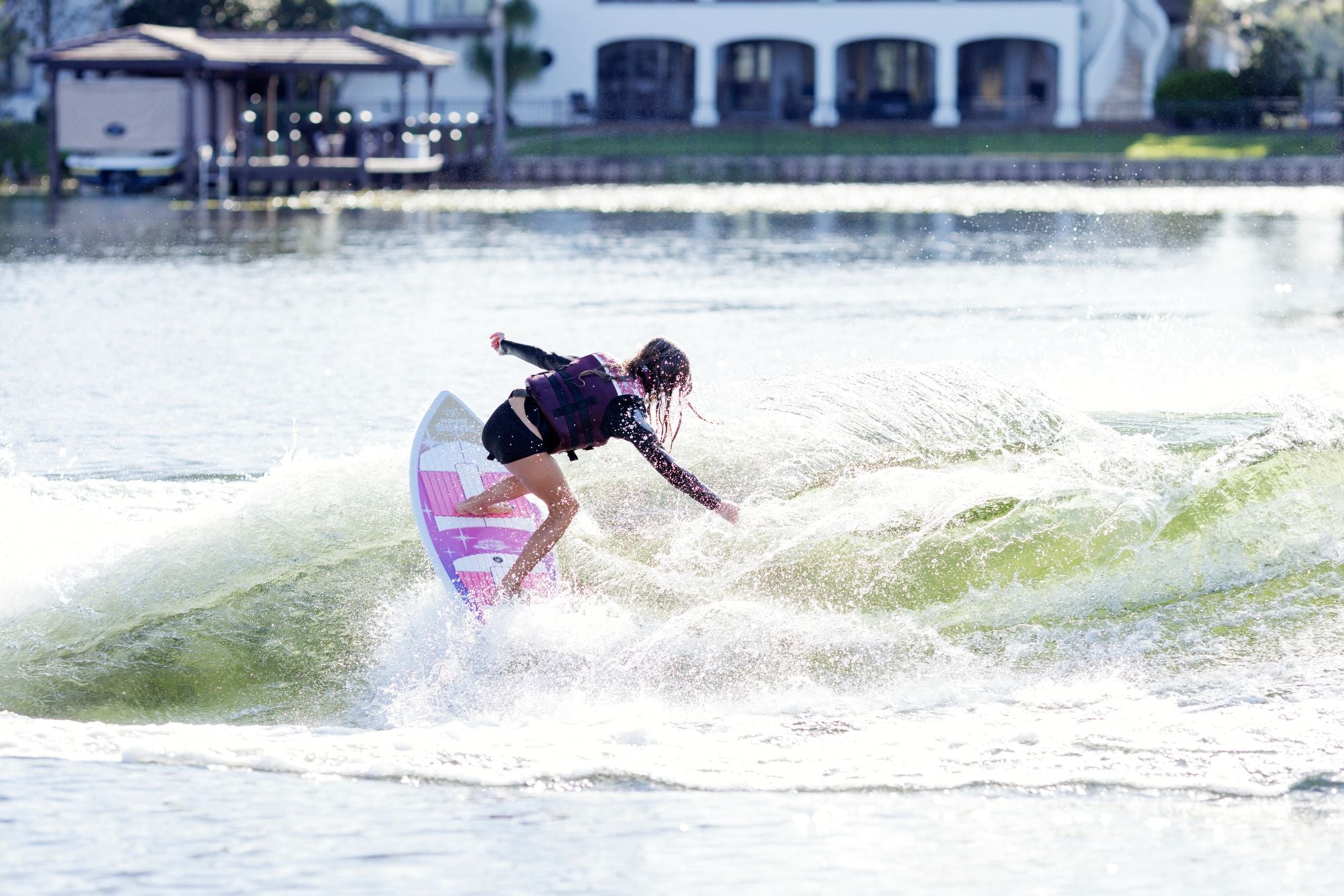 A person in a life vest rides the Ronix Sonic Girl's Fish Wakesurf Board 2026 - 3'9" on a sunlit lake, carving a spray of water while wake surfing with houses and docks in the background.