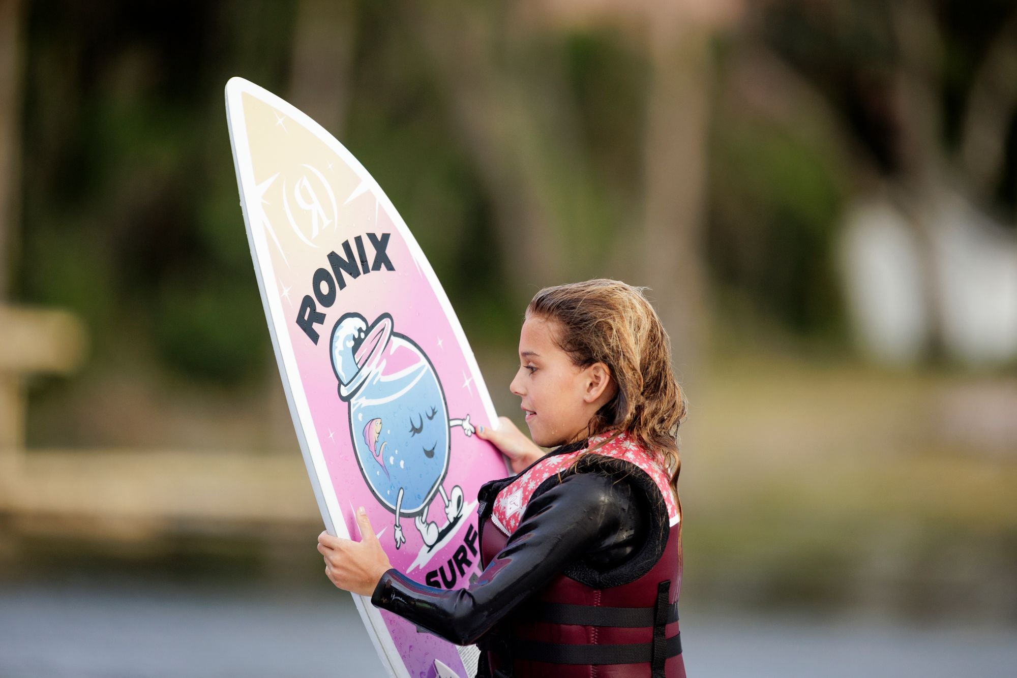 Wearing a life jacket, a young surfer holds the Ronix Sonic Girl's Fish Wakesurf Board 2026 (3'9")—a colorful board with cartoon graphics—standing by the water, ready to wake surf.