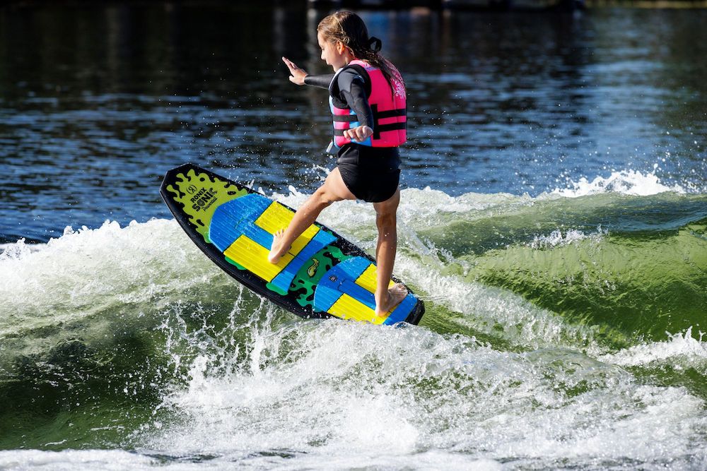 A girl in a pink life jacket and wetsuit surfs on a Ronix Sonic Powertail Wakesurf Board 2026 - 3'9", skillfully balancing on the colorful board as she rides a sunlit wave.