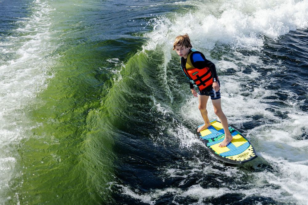 A young boy in a life jacket rides the Ronix Sonic Powertail Wakesurf Board 2026 - 3'9", gliding over greenish-blue water in bright sunlight.