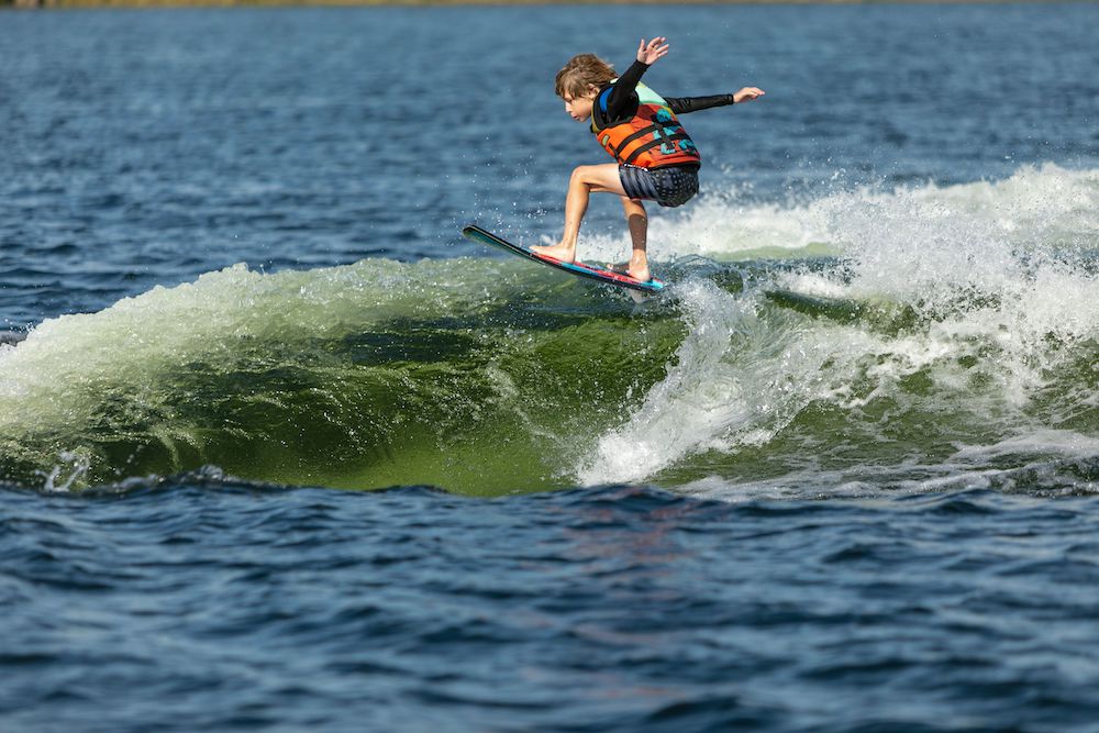 A young rider in a life jacket and shorts catches air above the wake on a Ronix Sonic Skimmer Wakesurf Board 2026 (3'11"), arms outstretched for balance over a large body of water.