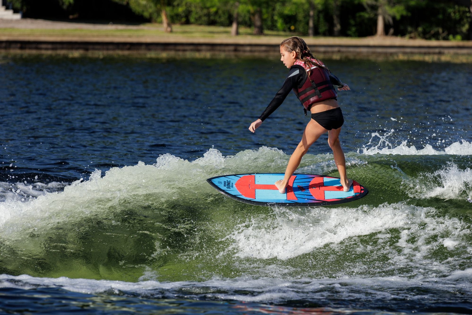 A young girl in a life vest rides the Ronix Sonic Skimmer Wakesurf Board 2026 (3'11") across a small wave, with trees and grass in the background.