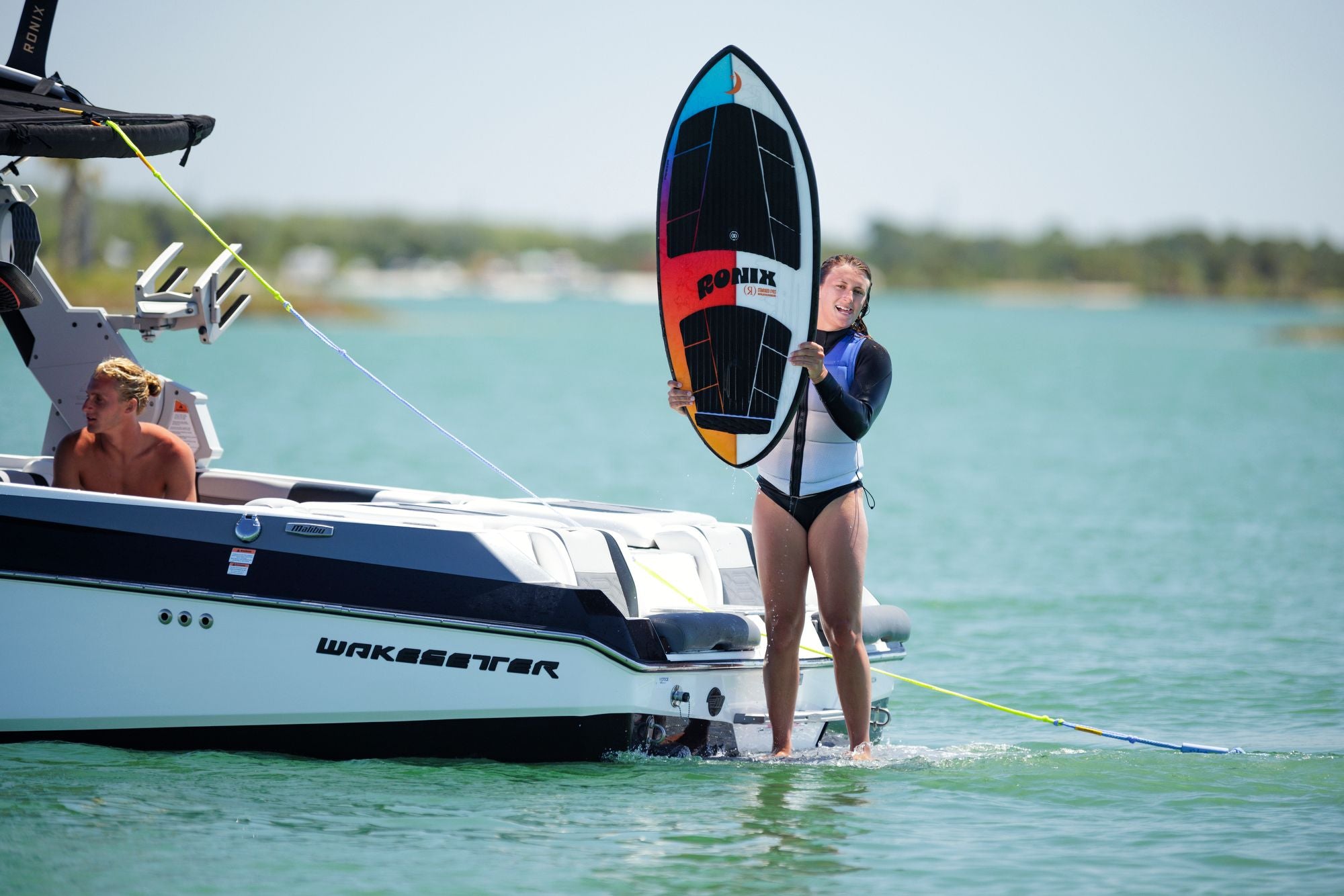 A woman in a wetsuit stands in shallow water beside a Wakesetter boat, holding a Ronix Standard Core Skimmer 2026—an ideal wakesurf board for beginner to intermediate riders. She smiles as the calm water mirrors the clear sky.