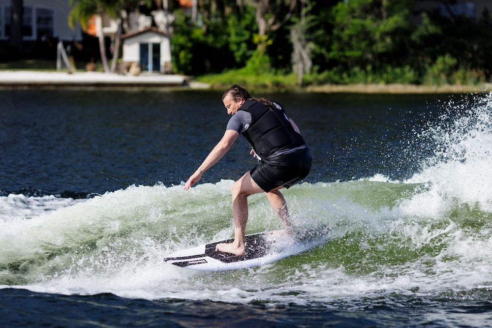 Wearing a black life vest and shorts, a surfer balances with style on a Ronix Wave King XL Surfer Wakesurf Board 2026, gliding across a river or lake surrounded by houses, trees, and lush greenery.