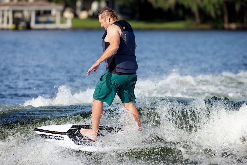 A man in a life vest and green shorts rides the Ronix Wave King XL Surfer Wakesurf Board 2026 by Ronix on a lake, generating small waves. Trees and a boathouse appear blurred in the background.