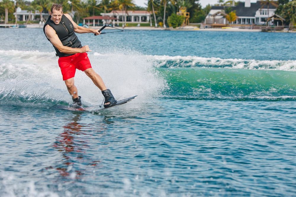 Wearing a Ronix Blackjack Yes Mens CGA Vest, a man in red shorts smiles as he wakeboards across a blue lake, holding a tow rope with water splashing behind him and houses and trees in the background.