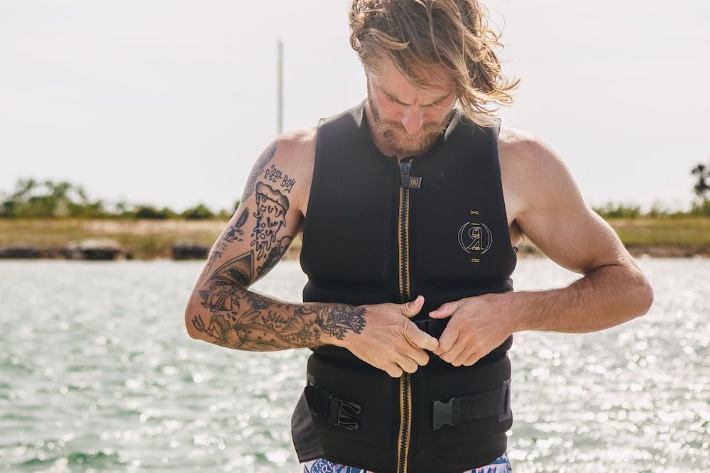 A tattooed man fastens a Ronix Blackjack Yes Mens CGA Vest by Ronix near water on a sunny day, with trees and grass in the background.