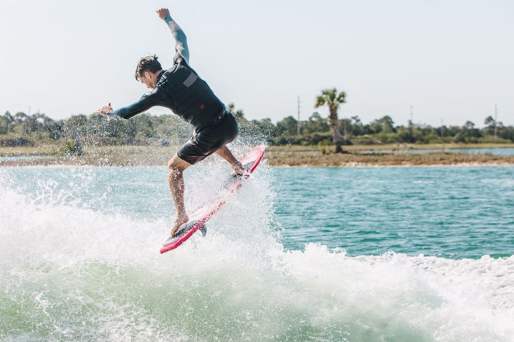 A competition-ready surfer in a black wetsuit rides a wave on the Ronix Flyweight Pro DNA Wakesurf Board 2026, performing a jump above the water on a sunny day with shoreline and palm trees in the background.