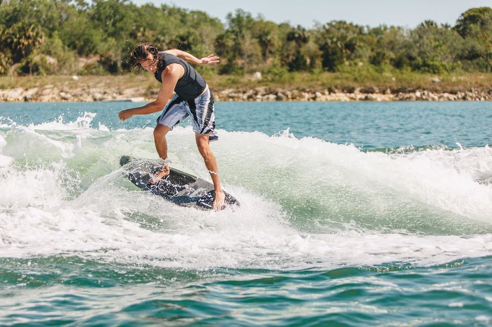 A person in a black life vest and striped shorts is wakeboarding on a sunny day, skillfully riding a Ronix M50 Dark Matter Wakesurf Board 2026 over a blue lake wave, with trees and rocks in the background.