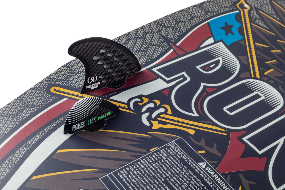 Close-up of the Ronix M50 Dark Matter Wakesurf Board 2026 with two fins—one black carbon Dark Matter fin and a green-and-black fin. The board displays a colorful graphic with “RONIX” and an eagle illustration.