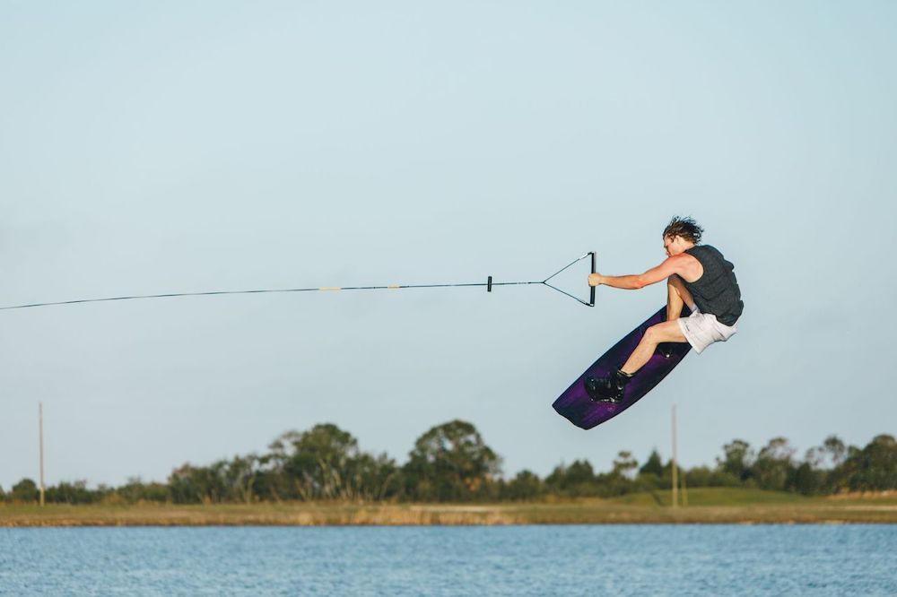 Wearing a life vest and Ronix One Gloss Midnight Boots 2026, a wakeboarder jumps high above the water, holding a tow rope, with trees and a clear sky in the background.