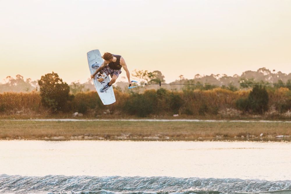 A wakeboarder wearing Ronix RXT Boots 2026 performs an aerial trick over the water at sunset while holding a tow rope, with trees and grassy land in the background.