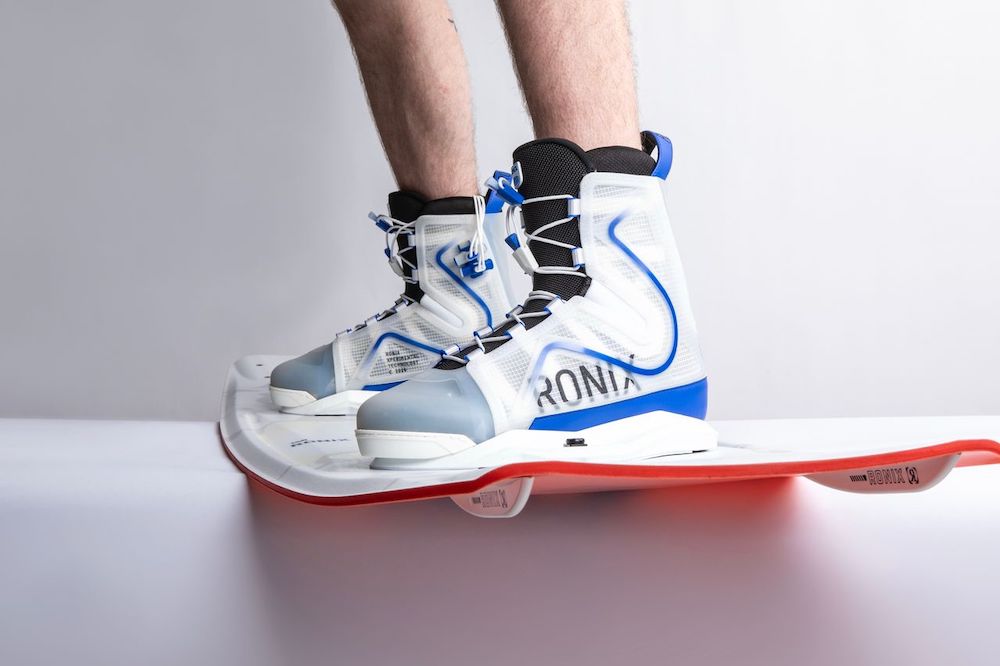Close-up of lower legs wearing Ronix RXT Boots 2026, white and blue boots by Ronix, attached to a white and red wakeboard against a plain background.