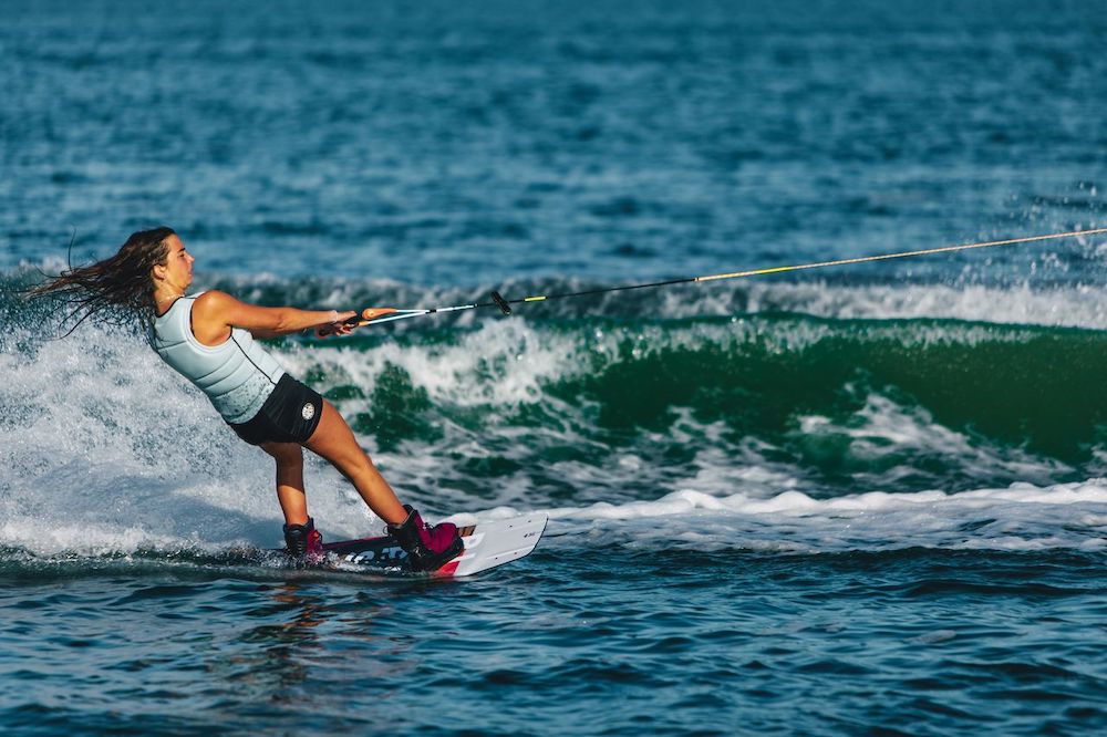 A woman wearing the Ronix Rise Women's CE Impact Vest 2026 from Ronix wakeboards across blue water, gripping a tow rope as waves and splashes follow behind.