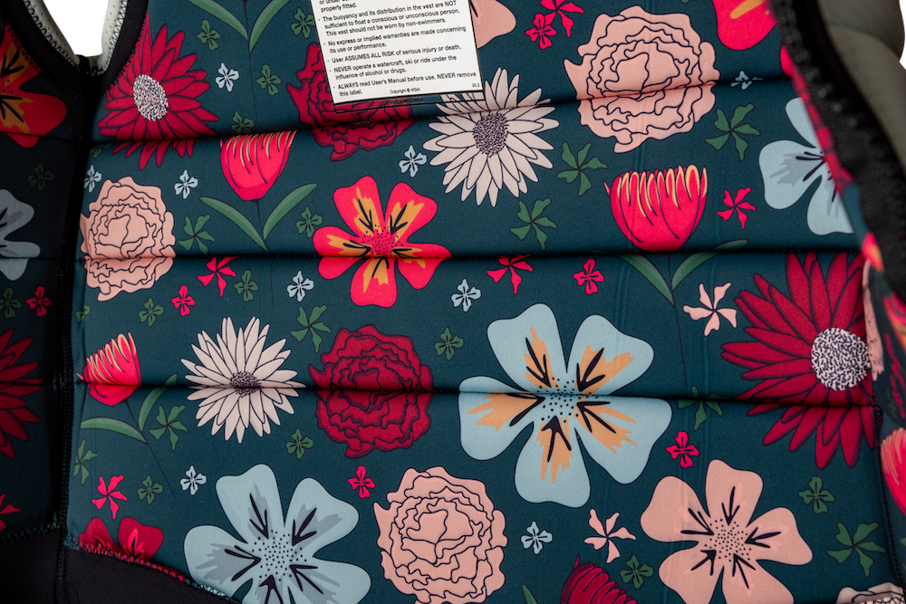 Close-up of the Ronix Rise Women's CE Impact Vest 2026 by Ronix, featuring a vibrant floral pattern with red, pink, yellow, and blue flowers and green leaves on a dark background; a white tag with printed text is visible at the top.