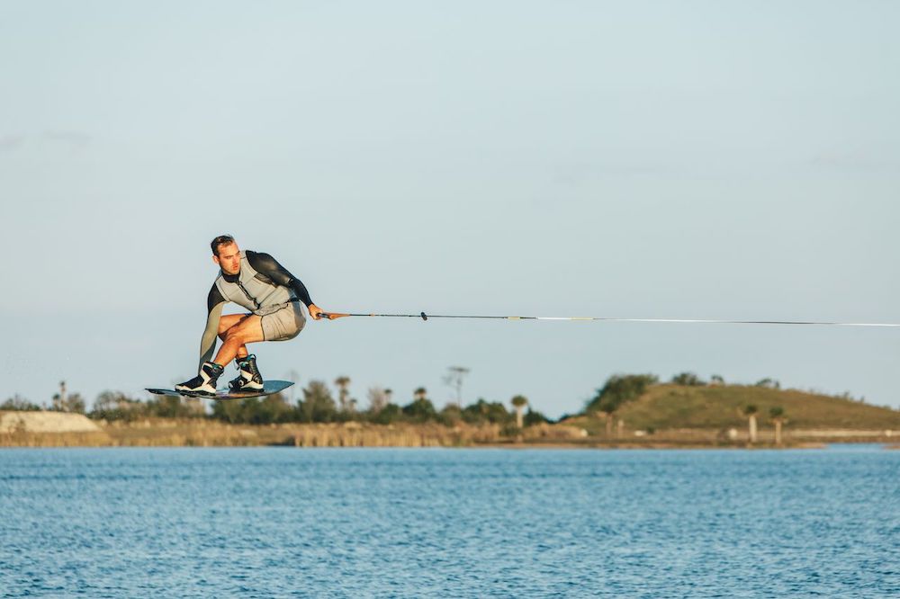 A man in a wetsuit is wakeboarding, gliding above the water while holding a rope, with grassy shoreline and blue sky in the background, showcasing the 2026 Ronix Supreme BOA Boots by Ronix.