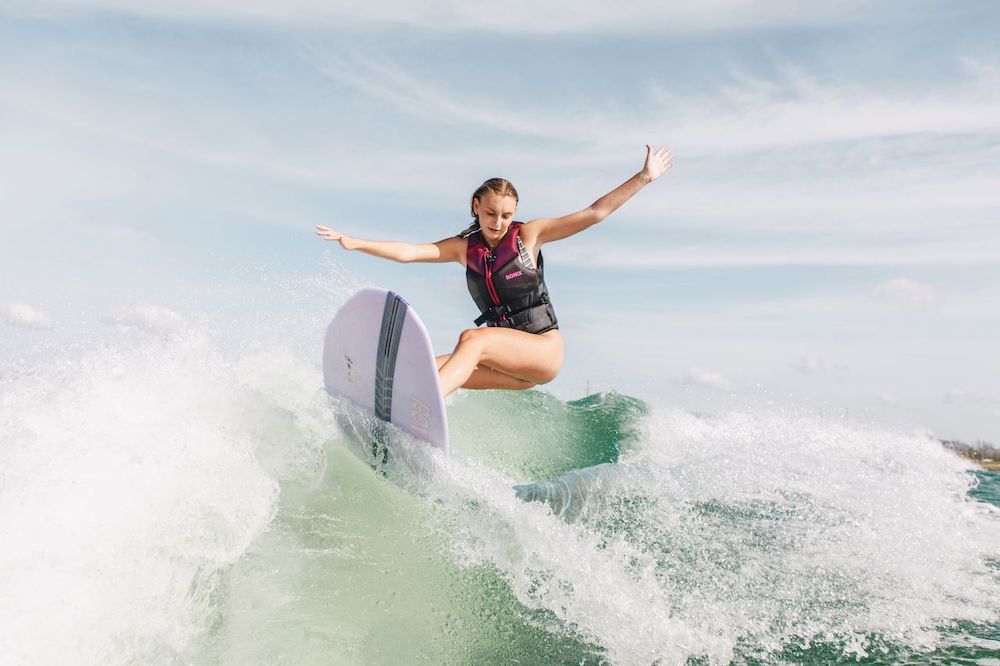 A woman wearing a life vest surfs on a wave, balancing with outstretched arms atop the Ronix Women's Aura Flyweight Skimmer 2026, against a backdrop of sky and ocean.
