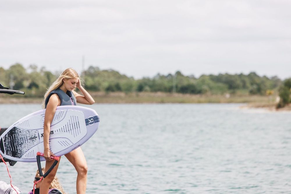 A woman in a life vest stands by the water holding the Ronix Women's Aura Flyweight Skimmer 2026, with trees and a cloudy sky in the background.