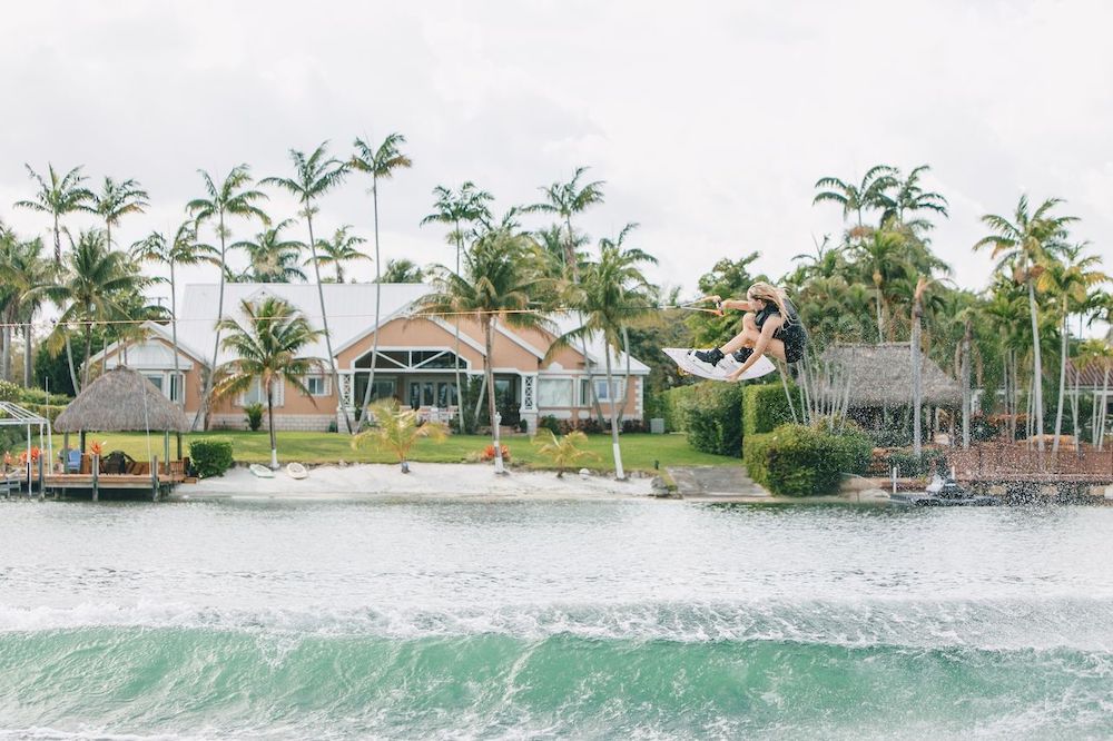 Someone rides the Ronix Quarter 'Til Midnight Wakeboard with Halo Bindings 2026, jumping above the water on a rope in front of a beach house with palm trees.