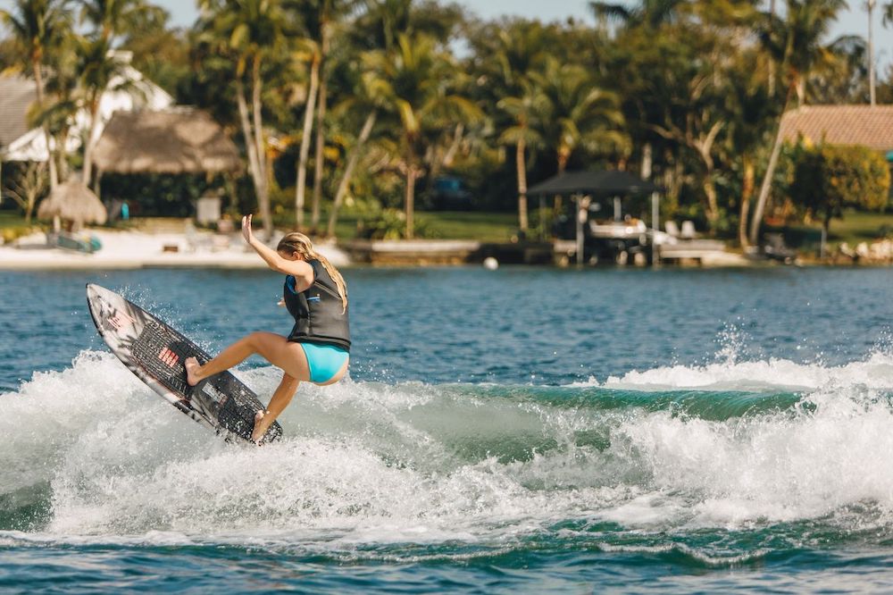 A woman rides the Ronix Women's Sea Captain Wakesurf Board 2026 on a sunny wave, wearing blue swimwear and a life vest, with palm trees and houses in the tropical background.