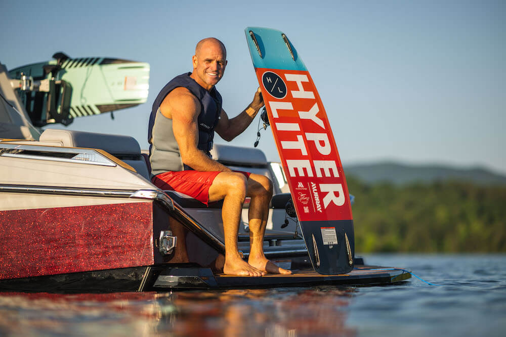 A man sitting on the back of a boat, holding a Hyperlite 2024 Murray Pro Wakeboard with a Subtle 3-Stage Rocker and Biolite 3 Core.