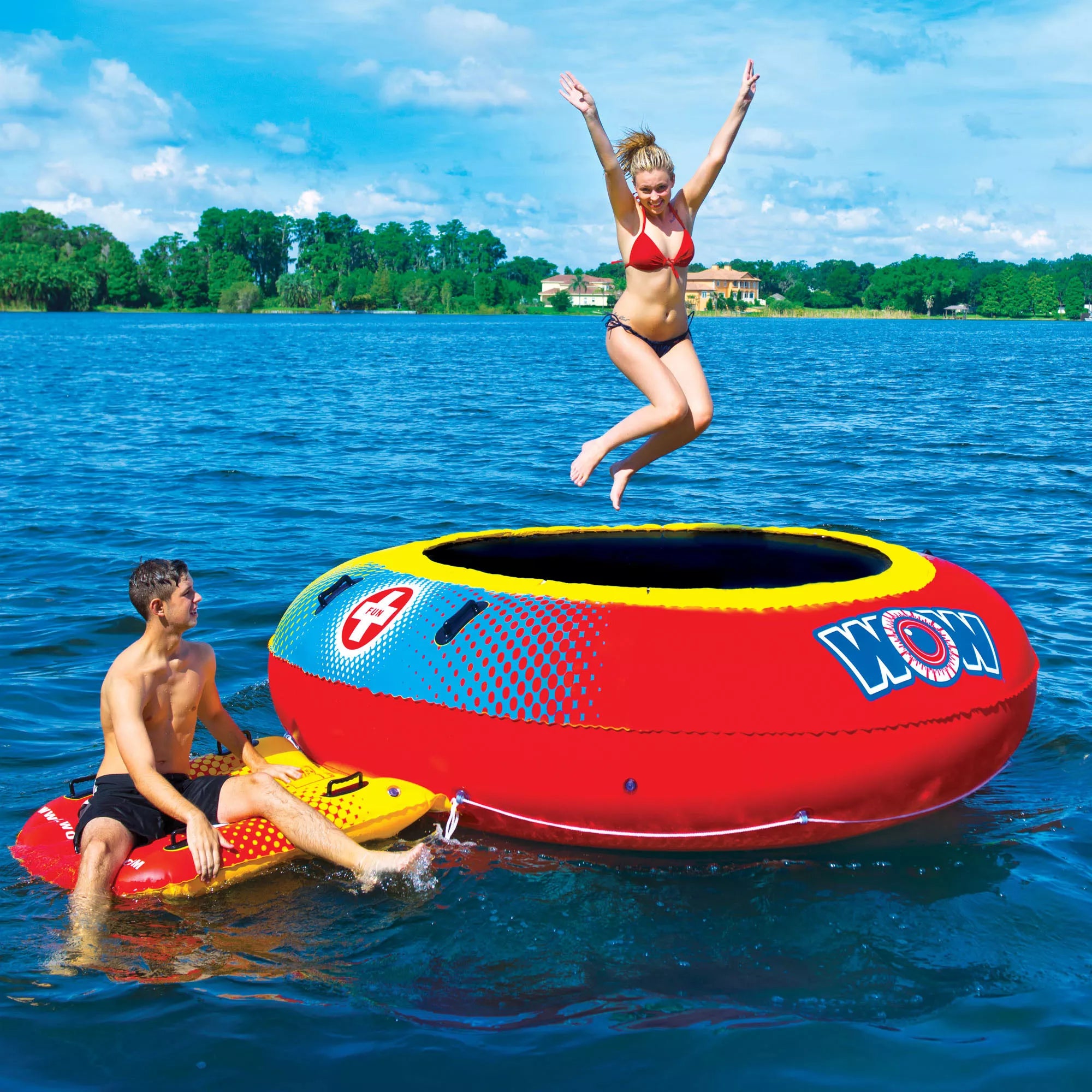A woman in a red bikini jumps onto the WOW 10' Bouncer by WOW on the lake, as a man in swim trunks relaxes on a smaller float nearby. Blue skies, trees, and lakeside houses complete the vibrant summer scene.