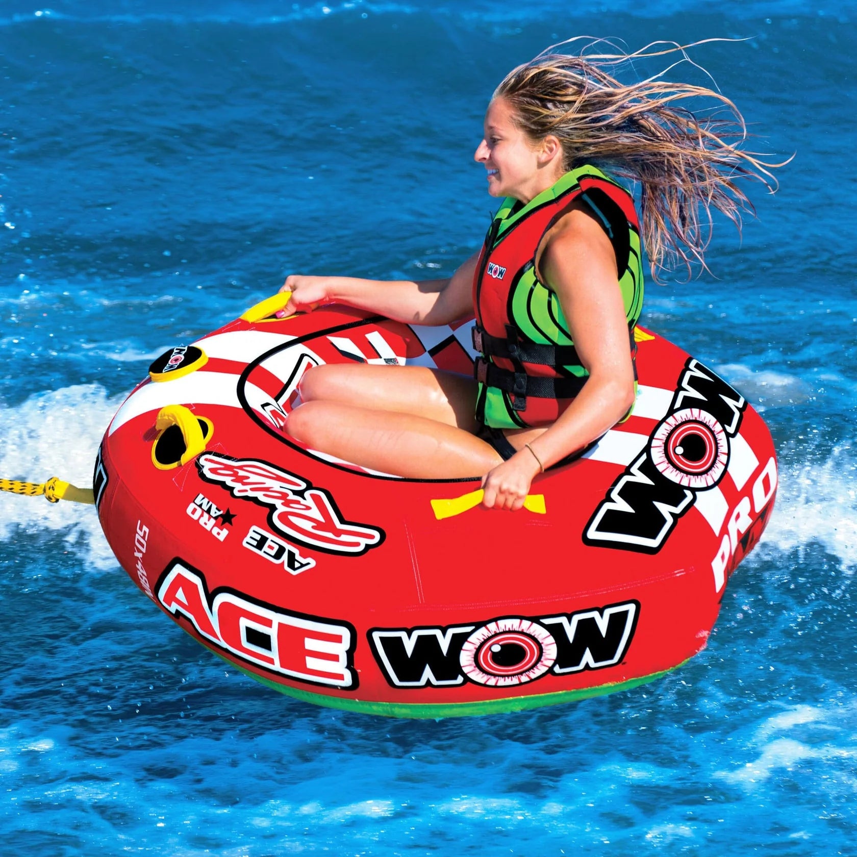 A woman in a life jacket smiles as she bounces over blue water on the WOW Ace Racer 1 Person Tube, a red and black boat tube with an arrow design and the WOW logo.