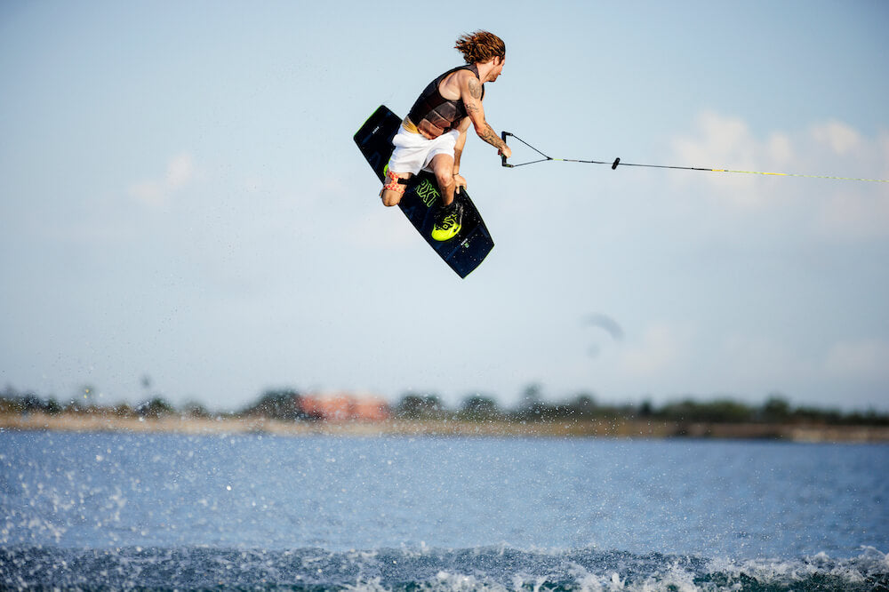 A person with long hair is wakeboarding on the Ronix 2024 RXT Blackout Wakeboard, catching air above the water while gripping a tow rope, enjoying ultra-soft landings under a clear blue sky with the shoreline in view.