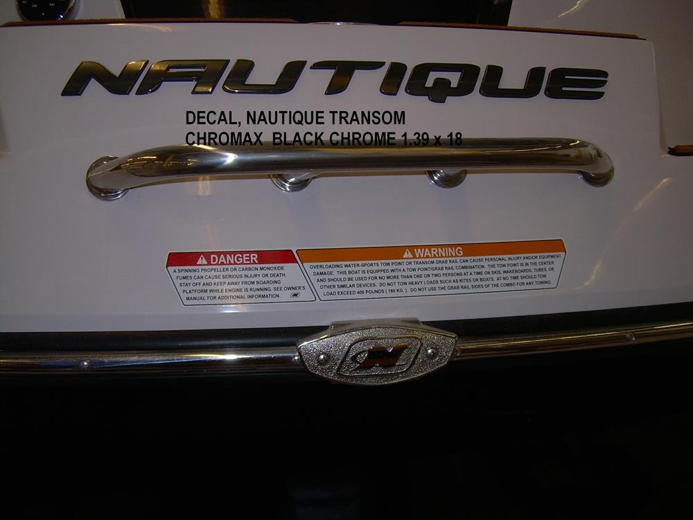 Close-up of a Nautique boat’s rear showing a metallic rail, warning labels, and a black chrome Nautique DECAL TRANSOM CHROMAX 1.39 x 18 (140017) above the transom.