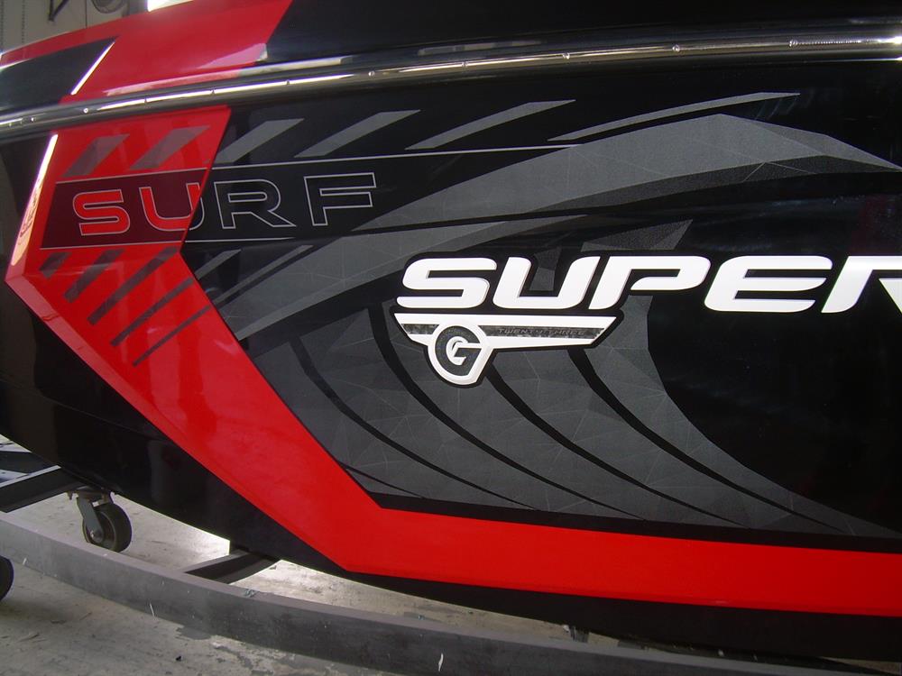 Close-up of the Nautique DECAL SURF GRAPHIC SET PORT & STARDBOARD G23 - 170353, featuring bold red, black, and gray graphics with “SURF,” “SUPER,” and a G23 Nautique decal, displayed on a boat resting on an indoor trailer.