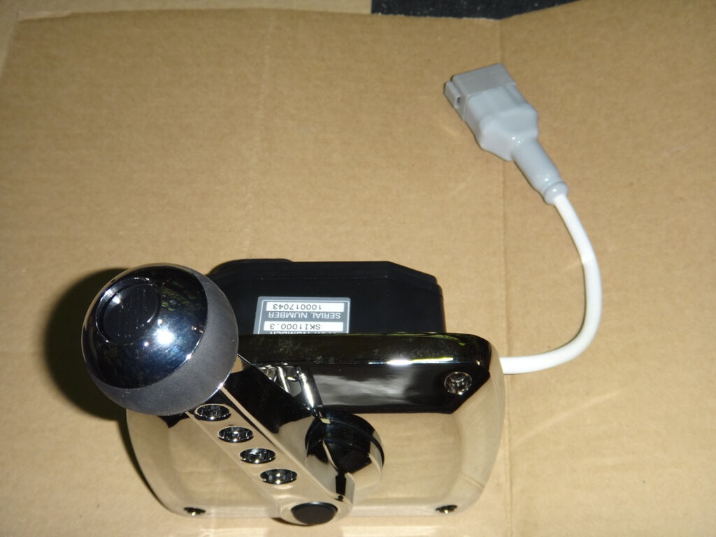 A close-up of a Malibu ZF Replacement Shifter E-Control 09-13 shows its shiny metallic lever with multiple black buttons, attached to a rectangular black and silver base, and a white cable with a gray connector, resting on cardboard.