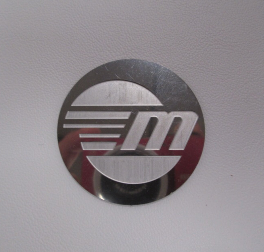 The Malibu Logo Steering Wheel Emblem features a metallic, circular design with a stylized "M" and horizontal lines on a white background. A faint photographer’s reflection appears in the emblem. Brand: Malibu.