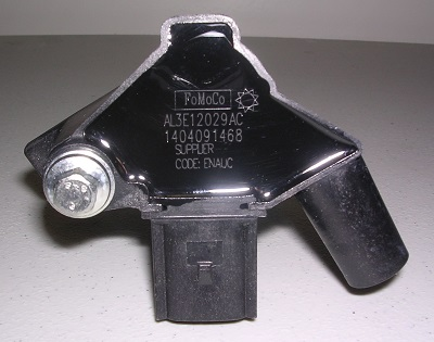 An Indmar COIL FORD 6.2 BANK 1 - 597031 automotive sensor, black with a bolt on the left and an electrical connector at the bottom, is shown on a gray surface. The brand is Indmar.