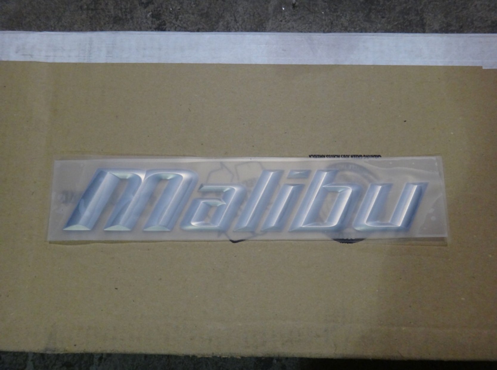 A Malibu SL24 Chrome Decal by Malibu is displayed on cardboard, with a concrete floor seen in the background.