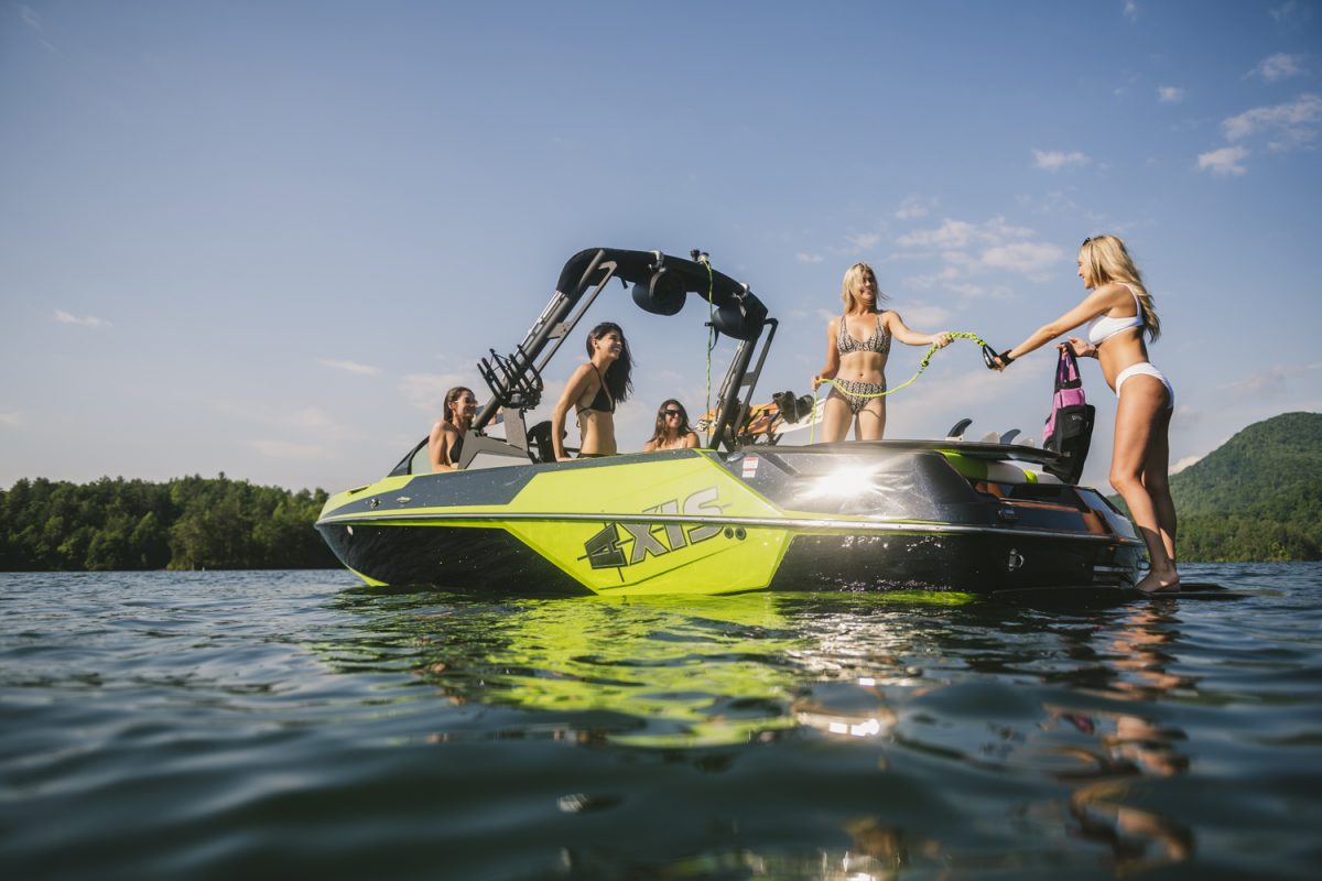 A group of young women in swimsuits relax and chat on a bright green speedboat featuring an Axis A-Emblem SL24-A decal, drifting on a calm lake with scenic trees and hills in the background under a sunny sky.