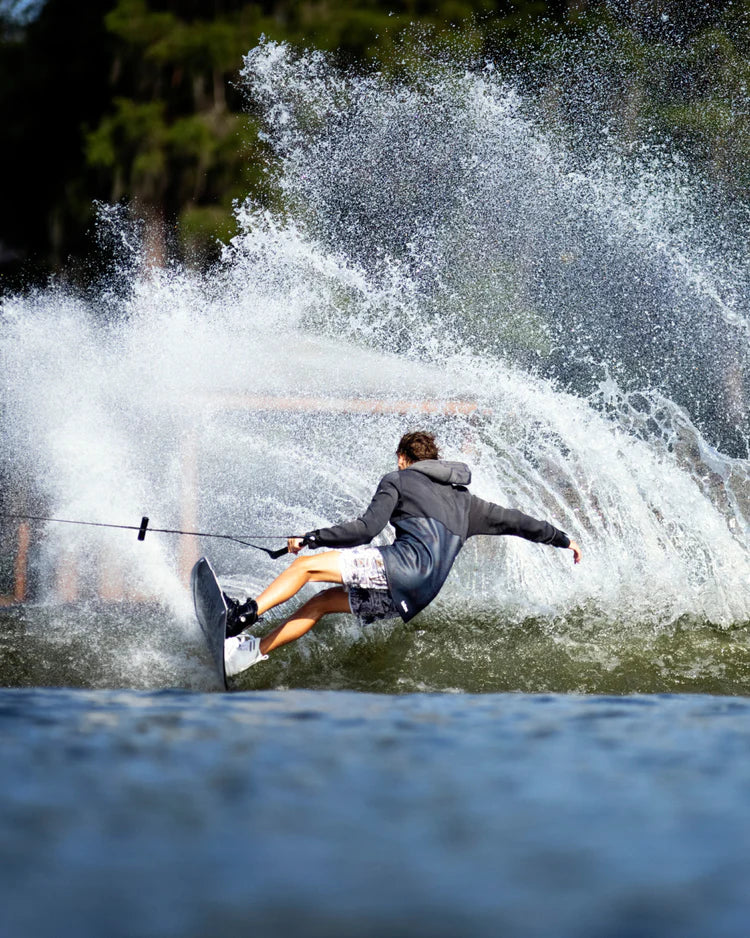 A man is riding a wakeboard on a lake, braving the wind chill factor in his Follow Wake 3.12 Anorak Pro Neo Outer Jacket - Charcoal.