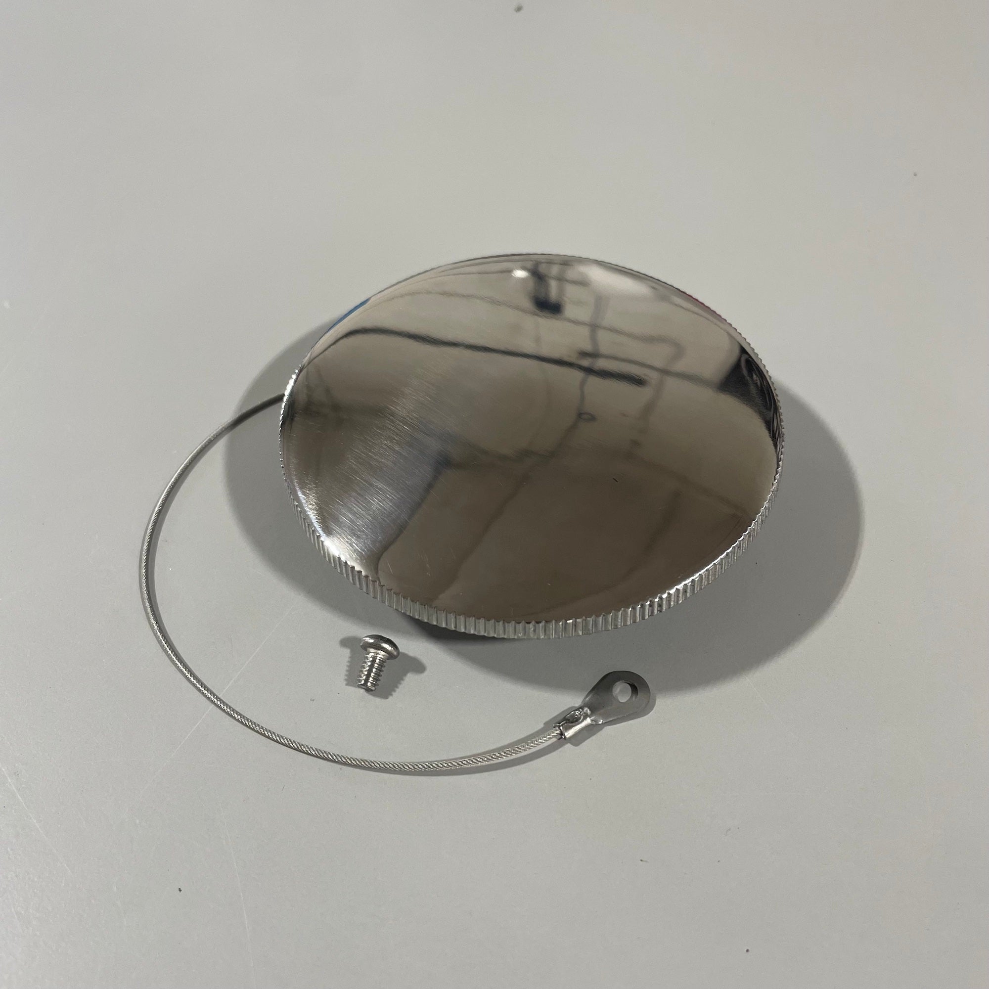 A shiny, round Malibu Gas Cap 9-11 (Replacement, Large Cap and Cable) rests on a light surface, accompanied by a small screw and thin attachment cable, each casting faint shadows.