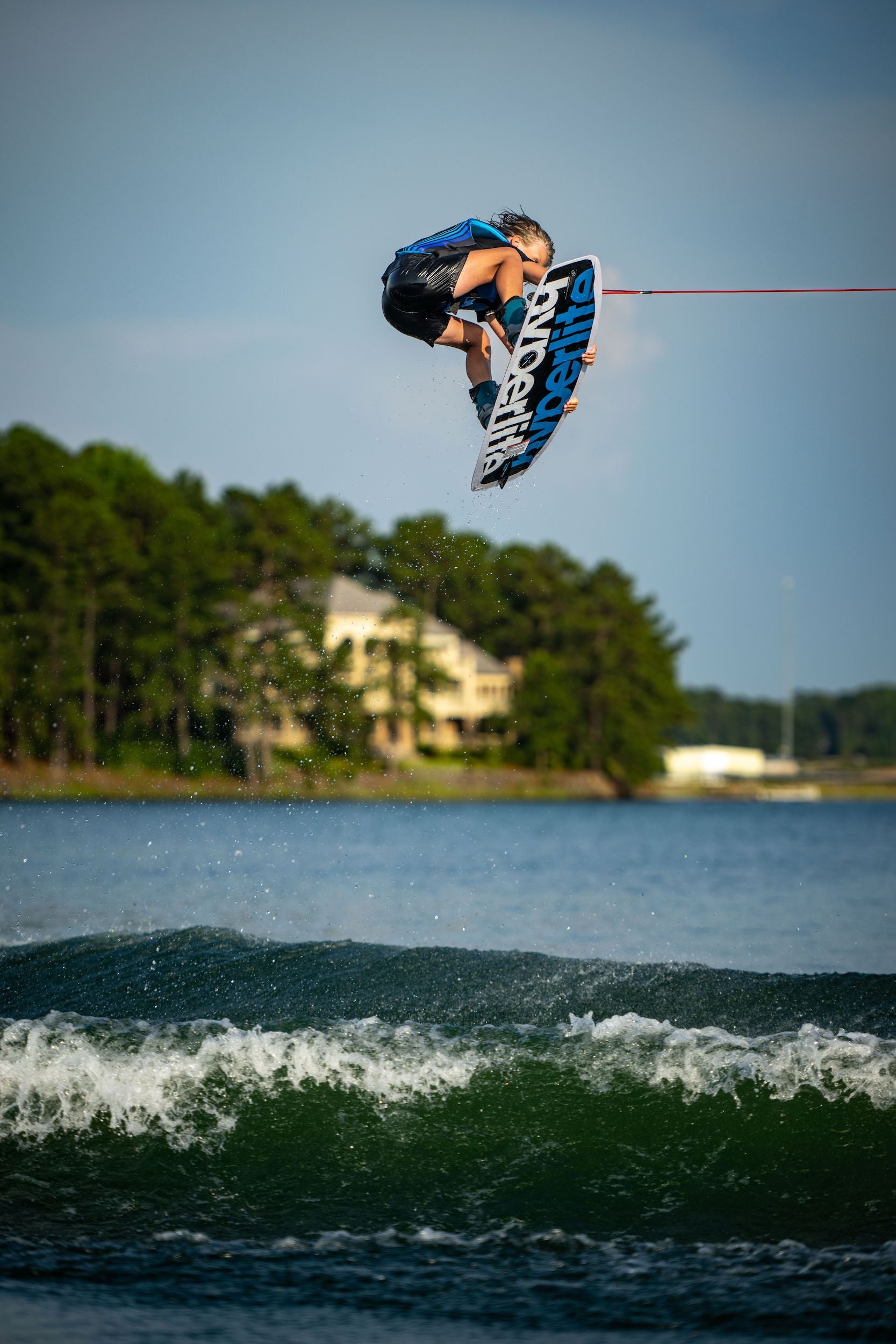 A beginner wakeboarder in the air over a lake, enjoying their forgiving first ride of the Hyperlite 2024 Motive Jr. Wakeboard - 119 from the free ride series.