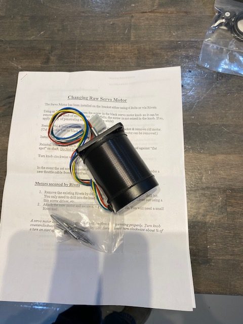 A black cylindrical Perfect Pass Raw Servo Motor (Motor Replacement, No Bracket, No Resistor) with colored wires sits on instructions and a wooden surface, with a bag of screws nearby for easy installation.