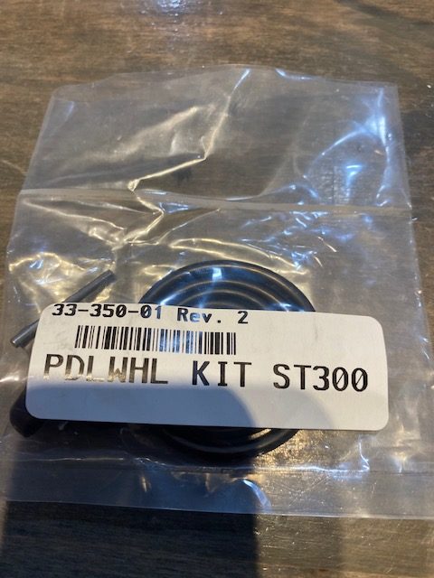 A sealed plastic bag labeled PDL WHL KIT ST300 holds a small metal pin and black circular part—components of the Perfect Pass Paddle Wheel Replacement Impeller & Shaft Kit (ST300)—from the brand Perfect Pass, on a wooden surface.