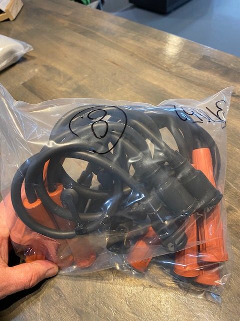 A hand holds a clear plastic bag with several Indmar Wire Spark Plug cables (Lower 6.2L Ford, part 597052) with orange connectors, placed on a wooden surface. Handwritten markings and numbers are visible on the bag.
