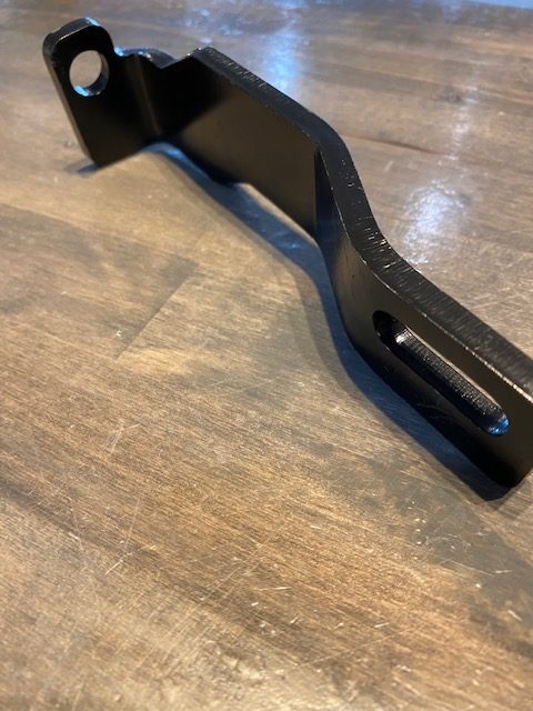 A close-up of the Indmar Bracket RWP Part "A" 6.2 Ford (985952), featuring a black, angled design with hole and slot ends, shown on a wooden surface. Designed by Indmar for use with 6.2 Ford engines.