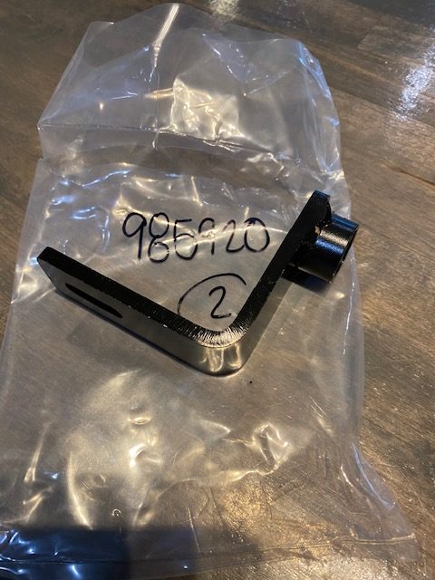 An Indmar Bracket Pump "B" Pump half 6.2 (985920), featuring a black metal L-shape with a cylindrical attachment, sits atop a sealed plastic bag labeled “98610” and “2” on a wooden surface.