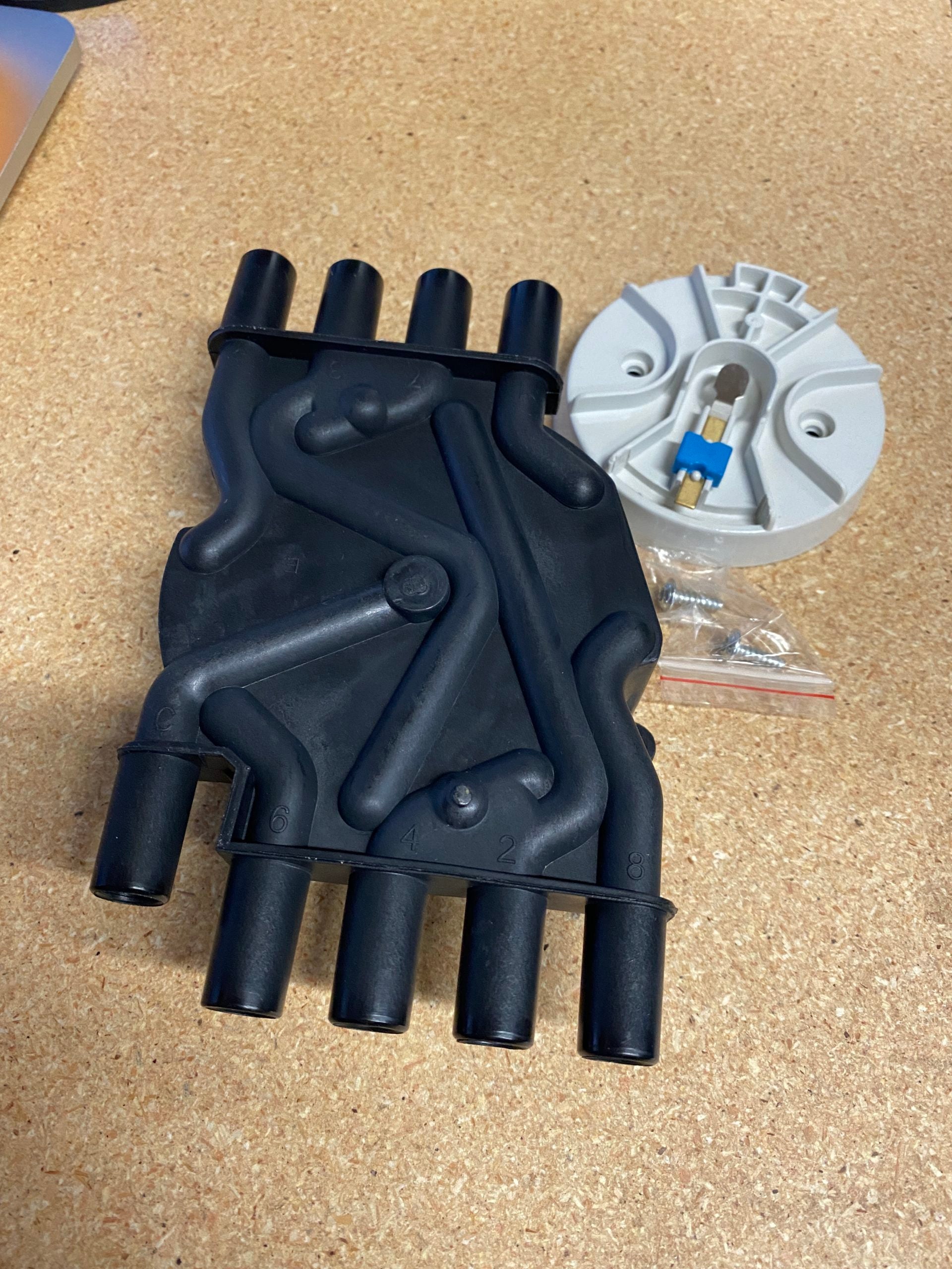 An Indmar Tune Kit: PCM ignition coil (black plastic, six terminals) lies beside a round white electrical part with a blue and gold connector, both set on a light wooden surface.