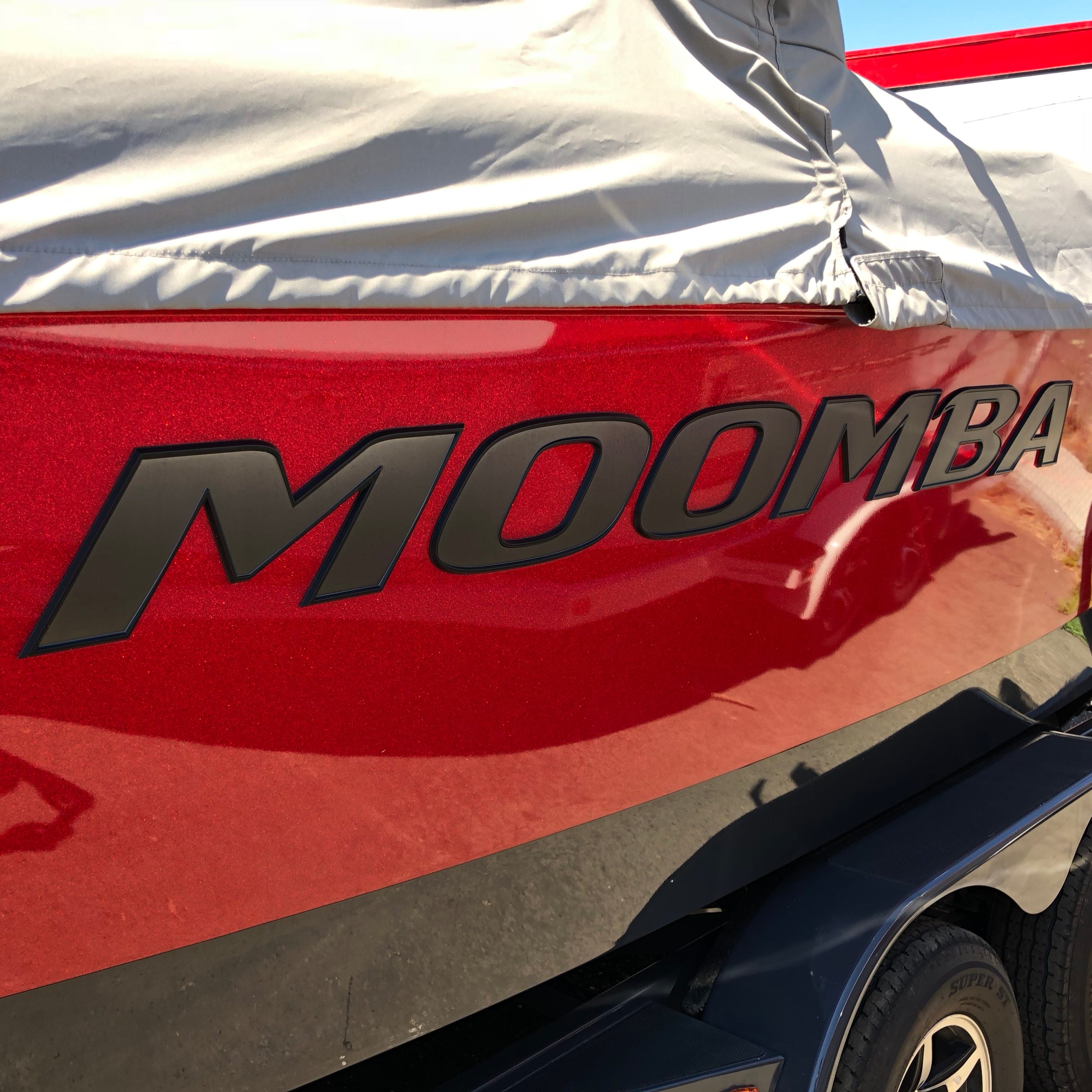 A close-up of a shiny red boat featuring a large Moomba Pro Black & Gun Metal Chromax (Hull Decal) by Moomba on the side, partially covered by a grey protective cover.