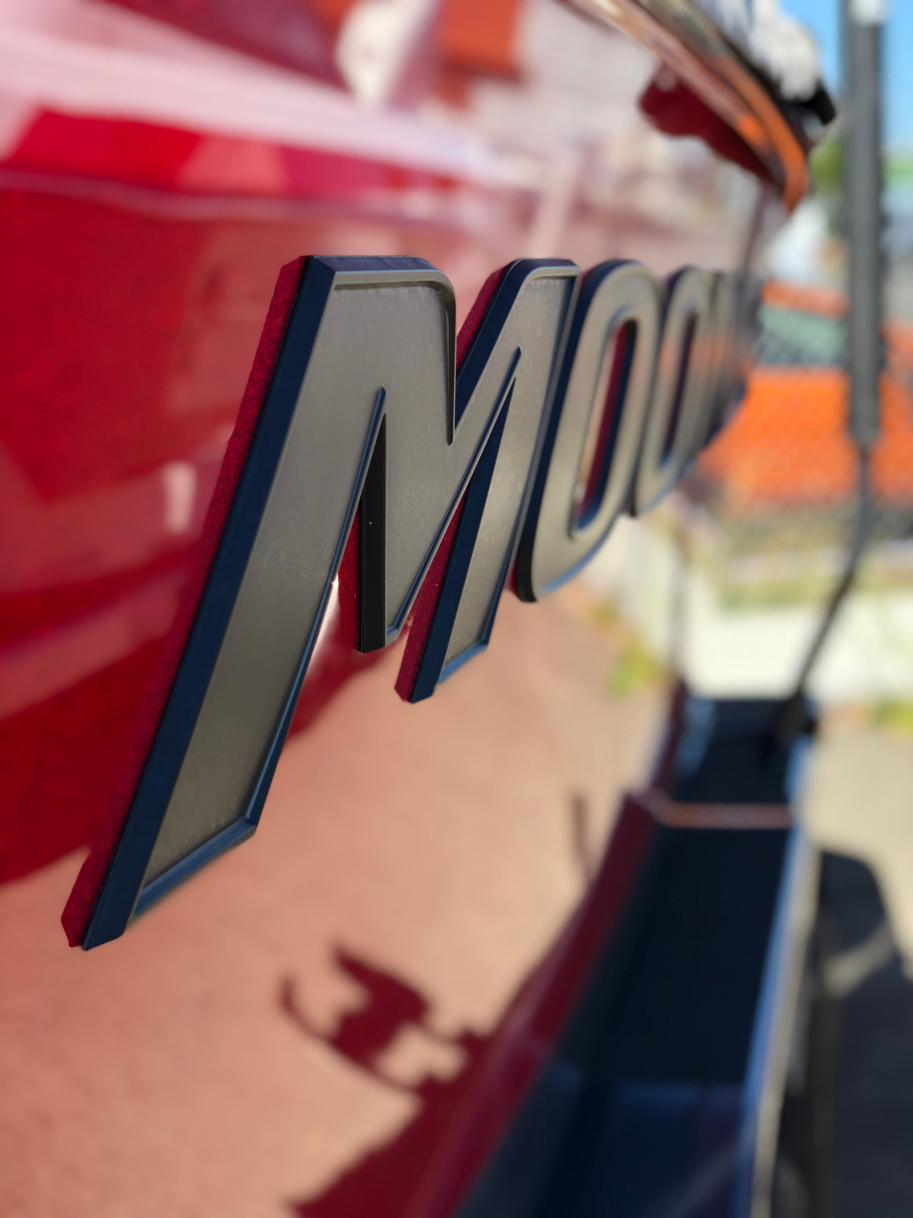 Close-up of bold, raised matte black "Moomba Pro Black & Gun Metal Chromax (Hull Decal)" letters by Moomba on a shiny red boat, with a blurred fence and outdoor background—showcasing this striking Moomba decal.